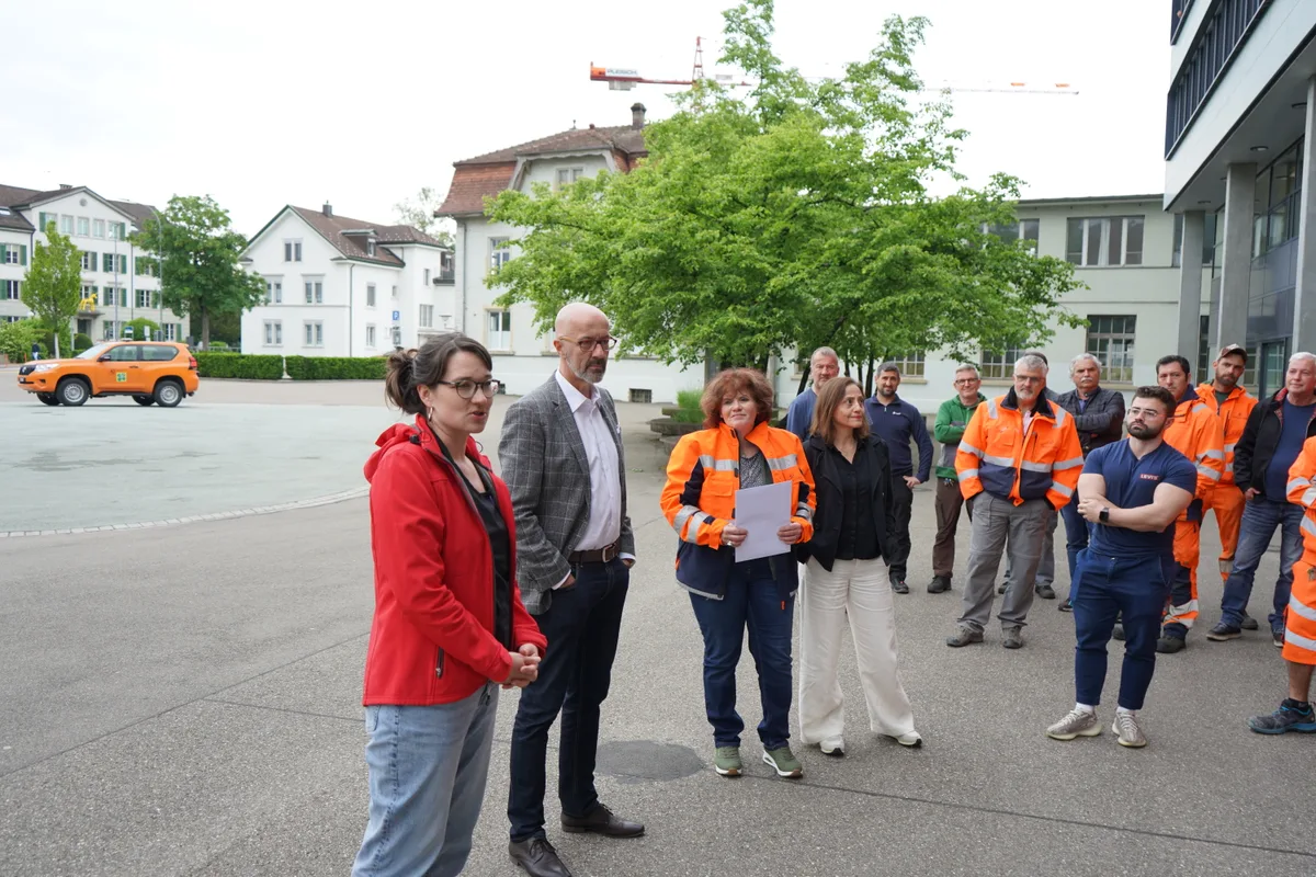Eine Gruppe von Leuten steht vor dem Stadthaus. Eine Frau mit einer roten Jacke hält eine Ansprache. 